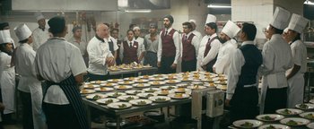 Movie still from “Hotel Mumbai” (2018), directed by Anthony Maras – A group of men standing in a kitchen with plates of food; Wide shot, High angle