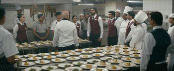 Movie still from “Hotel Mumbai” (2018), directed by Anthony Maras – A group of men standing in front of plates of food in a kitchen; Wide shot, High angle