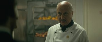 Movie still from “Hotel Mumbai” (2018), directed by Anthony Maras – An older man wearing a chef's outfit in front of a rack of fruit; Close Up shot, Over the shoulder angle