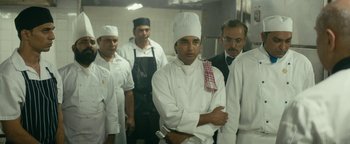 Movie still from “Hotel Mumbai” (2018), directed by Anthony Maras – A group of men in a kitchen wearing chef's hats and aprons; Medium shot, Over the shoulder angle