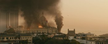 Movie still from “Hotel Mumbai” (2018), directed by Anthony Maras – A large building on fire with smoke billowing out of it; Extreme Wide shot, High angle