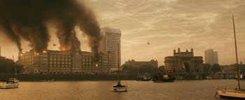 Movie still from “Hotel Mumbai” (2018), directed by Anthony Maras – A boat is in the water near a large building; Extreme Wide shot, High angle