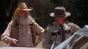 Movie still from “House of 1000 Corpses” (2003), directed by Rob Zombie – A man with a long beard standing next to a police officer; Medium shot, Over the shoulder angle