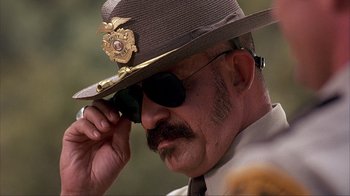 Movie still from “House of 1000 Corpses” (2003), directed by Rob Zombie – A police officer adjusts his hat while talking on a cell phone; Close Up shot, Low angle