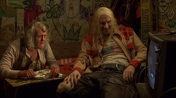 Movie still from “House of 1000 Corpses” (2003), directed by Rob Zombie – Two people sitting on a couch in front of a painting; Medium shot, Low angle