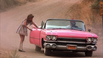 Movie still from “House of 1000 Corpses” (2003), directed by Rob Zombie – A woman standing next to a pink car on the side of a dirt road; Wide shot, High angle