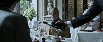 Movie still from “House of Gucci” (2021), directed by Ridley Scott – An older man sitting at a table with a wine bottle; Medium shot, Over the shoulder angle