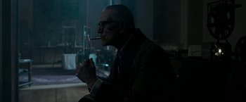 Movie still from “House of Gucci” (2021), directed by Ridley Scott – A man smoking a cigarette while sitting in a chair; Medium shot, Over the shoulder angle