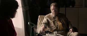 Movie still from “House of Gucci” (2021), directed by Ridley Scott – An old man sitting at a table smoking a cigarette; Medium shot, Over the shoulder angle