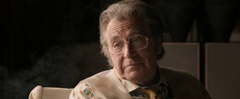 Movie still from “House of Gucci” (2021), directed by Ridley Scott – An older man wearing glasses and a striped shirt; Close Up shot, Over the shoulder angle