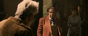 Movie still from “House of Gucci” (2021), directed by Ridley Scott – An older man wearing a suit and tie standing in front of two other people; Medium shot, Over the shoulder angle