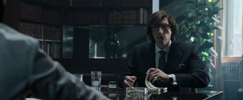 Movie still from “House of Gucci” (2021), directed by Ridley Scott – A man sitting at a wooden table in a room; Medium shot, Over the shoulder angle