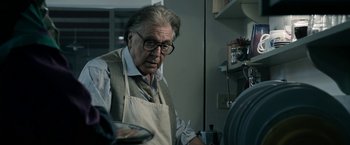 Movie still from “House of Gucci” (2021), directed by Ridley Scott – An older man wearing glasses and an apron in a kitchen; Close Up shot, Over the shoulder angle