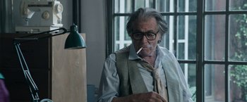 Movie still from “House of Gucci” (2021), directed by Ridley Scott – An older man smoking a cigarette while wearing glasses; Medium shot, Over the shoulder angle