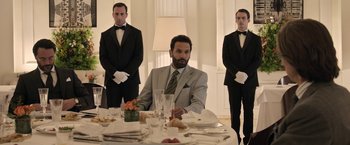 Movie still from “House of Gucci” (2021), directed by Ridley Scott – A man sitting at a dinner table in front of two other men in tuxedos; Medium shot, Over the shoulder angle