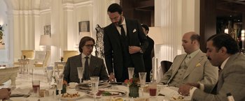 Movie still from “House of Gucci” (2021), directed by Ridley Scott – A man in a black suit standing at a dinner table; Medium shot, Over the shoulder angle