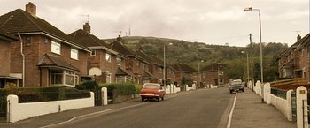 Movie still from “Hunger” (2008), directed by Steve McQueen – A car is driving down the street in front of a row of houses; Extreme Wide shot, High angle