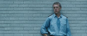 Movie still from “Hunger” (2008), directed by Steve McQueen – A police officer leaning against a brick wall; Medium shot, Low angle
