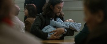 Movie still from “Hunger” (2008), directed by Steve McQueen – A man holding a baby in his arms while sitting at a table; Medium shot, Low angle