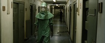 Movie still from “Hunger” (2008), directed by Steve McQueen – A man in a green suit and a mask walks down a hallway; Wide shot, High angle