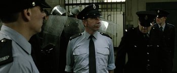 Movie still from “Hunger” (2008), directed by Steve McQueen – A man in a police uniform standing in front of a metal fence; Medium shot, Low angle