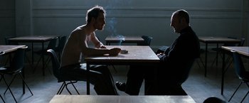 Movie still from “Hunger” (2008), directed by Steve McQueen – Two men sitting at a table with a cigarette in their mouth; Medium shot, Over the shoulder angle