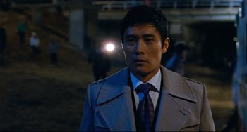 Movie still from “I Saw the Devil” (2010), directed by Jee-woon Kim – A man wearing a suit and tie standing in front of a building; Close Up shot, Over the shoulder angle