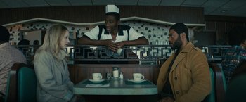 Movie still from “I'm Your Woman” (2020), directed by Julia Hart – Two people sitting at a table in front of a counter; Medium shot, Over the shoulder angle