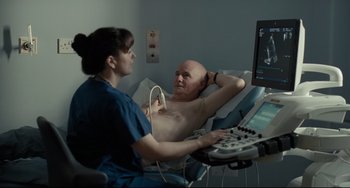 Movie still from “I, Daniel Blake” (2016), directed by Ken Loach – A woman is talking to a man who is laying on a bed; Medium shot, Over the shoulder angle