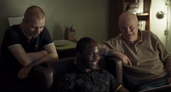 Movie still from “I, Daniel Blake” (2016), directed by Ken Loach – Three people sitting in a chair and one of them is smiling; Medium shot, Over the shoulder angle