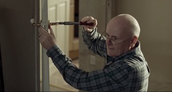 Movie still from “I, Daniel Blake” (2016), directed by Ken Loach – An older bald man in a plaid shirt using a screwdriver to fix a door; Medium shot, Over the shoulder angle