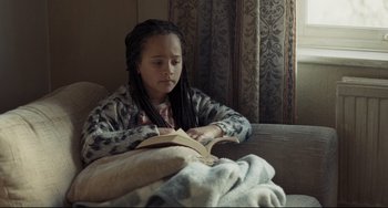 Movie still from “I, Daniel Blake” (2016), directed by Ken Loach – A girl sitting on a couch reading a book; Close Up shot, Over the shoulder angle