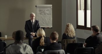 Movie still from “I, Daniel Blake” (2016), directed by Ken Loach – A man sitting in front of a group of people in front of a flip chart; Wide shot, Over the shoulder angle