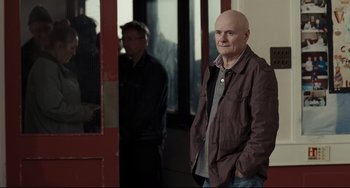 Movie still from “I, Daniel Blake” (2016), directed by Ken Loach – A man standing in front of a red door with another man standing behind him; Medium shot, Over the shoulder angle