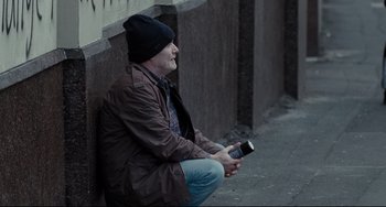 Movie still from “I, Daniel Blake” (2016), directed by Ken Loach – A man sitting on the side of the street holding a cell phone; Medium shot, Over the shoulder angle