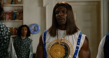 Movie still from “Idiocracy” (2006), directed by Mike Judge – A man in a white shirt with a medal around his neck; Medium shot, Low angle