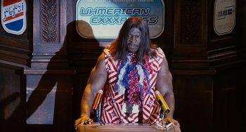 Movie still from “Idiocracy” (2006), directed by Mike Judge – A man dressed in a red , white , and blue shirt standing at a podium with microphones; Medium shot, Low angle