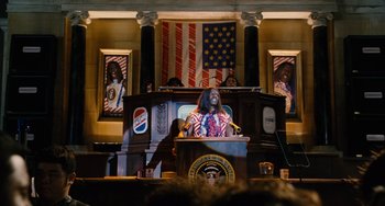 Movie still from “Idiocracy” (2006), directed by Mike Judge – A man is giving a speech in front of an american flag; Medium shot, Low angle