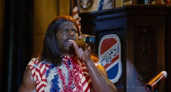 Movie still from “Idiocracy” (2006), directed by Mike Judge – A man with long black hair is holding a bottle of soda; Medium shot, Low angle