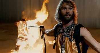 Movie still from “Idiocracy” (2006), directed by Mike Judge – A bearded man holding a hose and a fire in the background; Medium shot, Over the shoulder angle