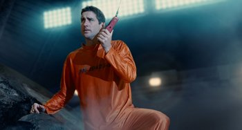 Movie still from “Idiocracy” (2006), directed by Mike Judge – A man holding a cell phone while standing in an arena; Medium shot, Low angle