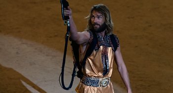 Movie still from “Idiocracy” (2006), directed by Mike Judge – A man in a gold outfit holding a black leash; Medium shot, Over the shoulder angle