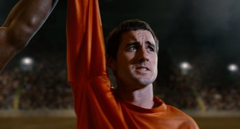 Movie still from “Idiocracy” (2006), directed by Mike Judge – A man in an orange shirt holding his arm up; Close Up shot, Low angle