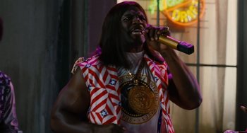 Movie still from “Idiocracy” (2006), directed by Mike Judge – A man with long black hair is holding a microphone and singing; Medium shot, Low angle
