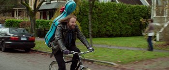 Movie still from “If I Stay” (2014), directed by R.J. Cutler – A man riding a bike with a large blue bag on the back of it; Medium shot, High angle