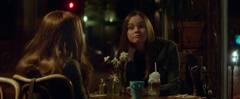 Movie still from “If I Stay” (2014), directed by R.J. Cutler – A woman sitting at a table in front of a vase of flowers; Medium shot, Over the shoulder angle
