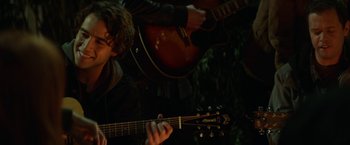 Movie still from “If I Stay” (2014), directed by R.J. Cutler – A man playing an acoustic guitar in a dark forest; Close Up shot, Over the shoulder angle