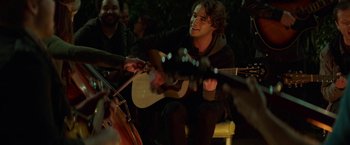 Movie still from “If I Stay” (2014), directed by R.J. Cutler – A man playing an acoustic guitar in front of a group of people; Medium shot, Over the shoulder angle