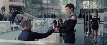 Movie still from “In Time” (2011), directed by Andrew Niccol – A woman in a black dress is handing a book to a man in a black dress; Medium shot, Over the shoulder angle