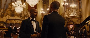 Movie still from “In Time” (2011), directed by Andrew Niccol – Two men in tuxedos talking in front of chandeliers; Medium shot, Over the shoulder angle
